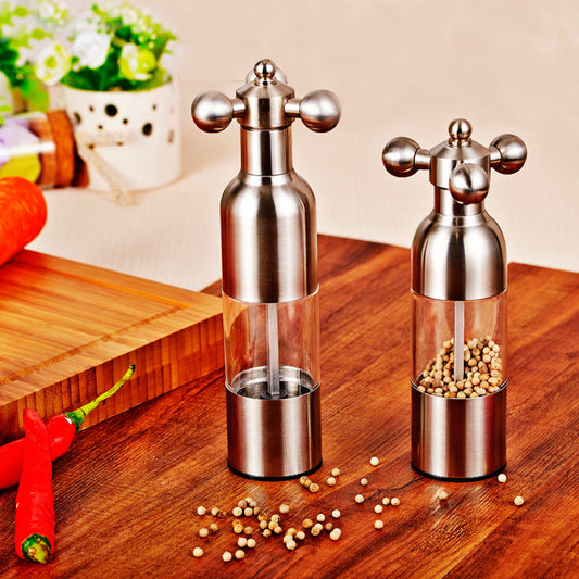 Two silver pepper mills on a wooden surface with spices around.