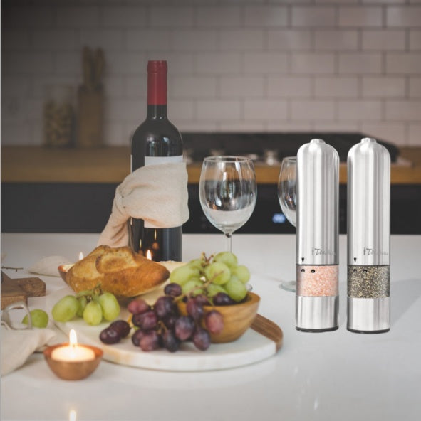 Two pepper mills on a table with a bottle of wine, glasses, and fruit platter.