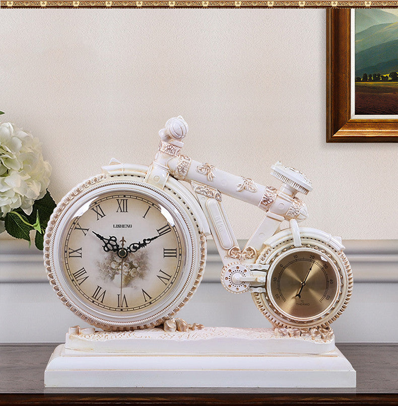 Decorative clock shaped like a bicycle on a table with a floral arrangement and painting in the background.