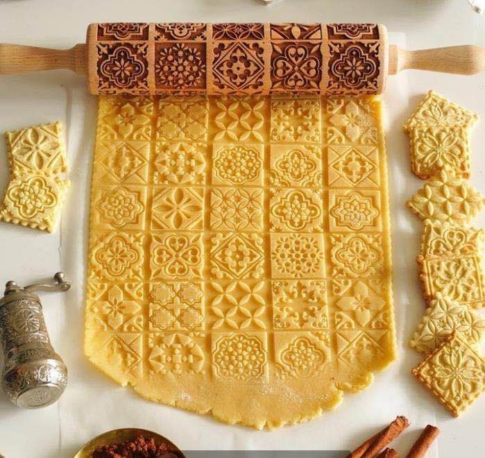 Decorative rolling pin with embossed patterns on rolled-out dough, surrounded by cookies and spices.