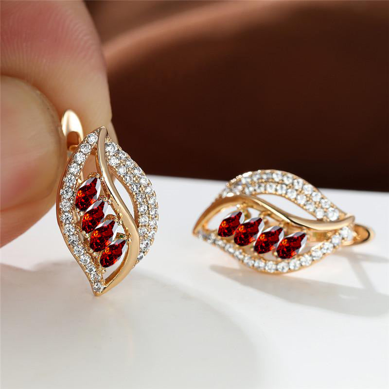 Gold earrings with red gemstones held by a hand on a white surface.
