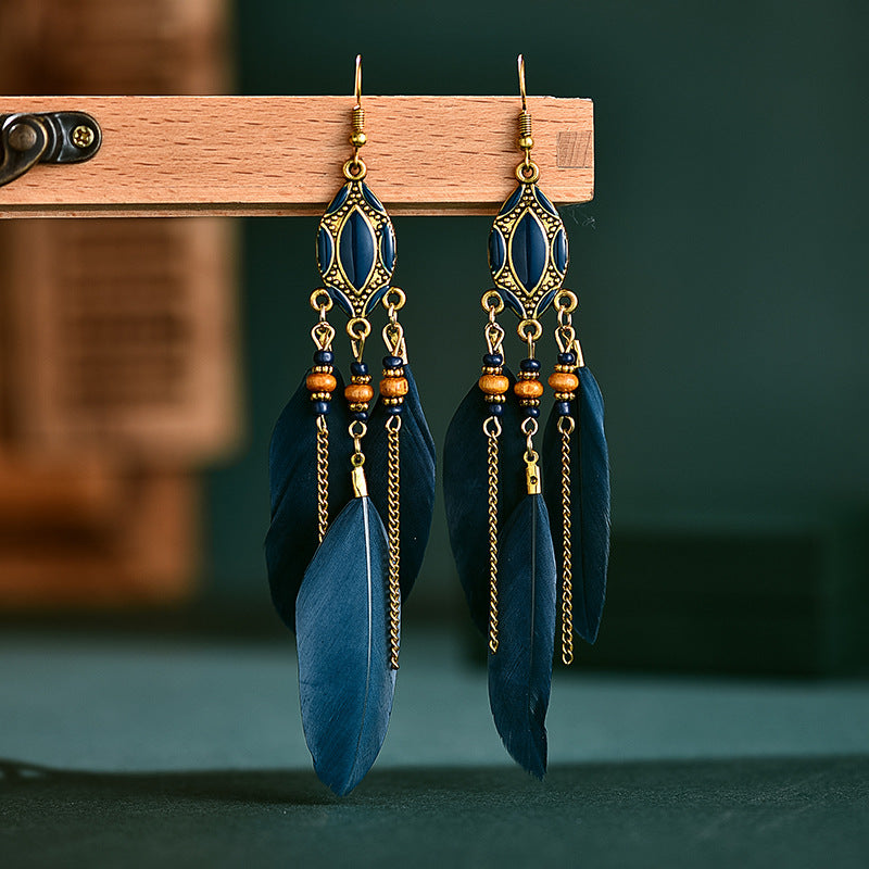 Blue feather earrings with gold chains hanging on a wooden stand against a dark green background. From Nested Wisdom