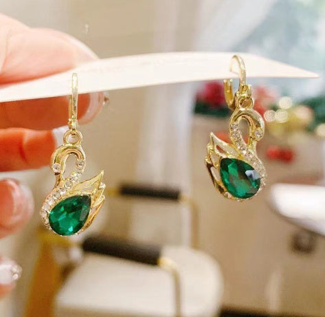 Gold Swan earrings with green gemstones held by a hand against a blurred background.