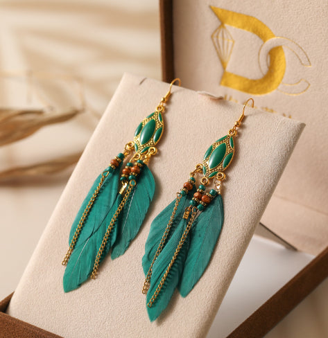 Turquoise feather earrings with gold accents on a beige stand. From Nested Wisdom