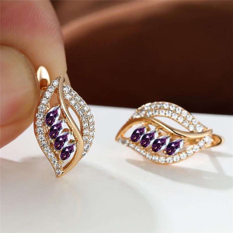 Gold earrings with purple gemstones held by a hand on a white surface.