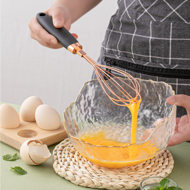 Person whisking eggs in a glass bowl with a copper whisk, surrounded by eggs and a cutting board on a light surface.