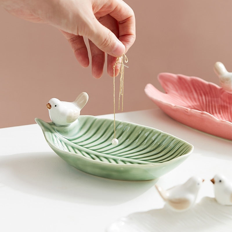 Hand placing a necklace on a green ceramic leaf-shaped dish with small bird figurines.